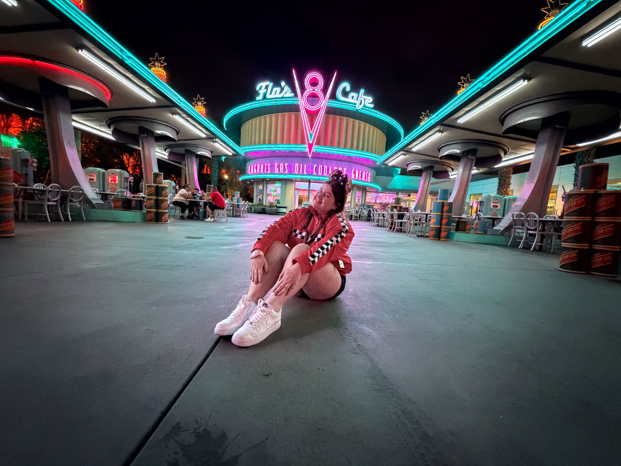 A guest in a red racing jacket and Minnie Mouse ears sits on the pavement in front of Flo's V8 Cafe in Cars Land at night, beneath its glowing neon signs.