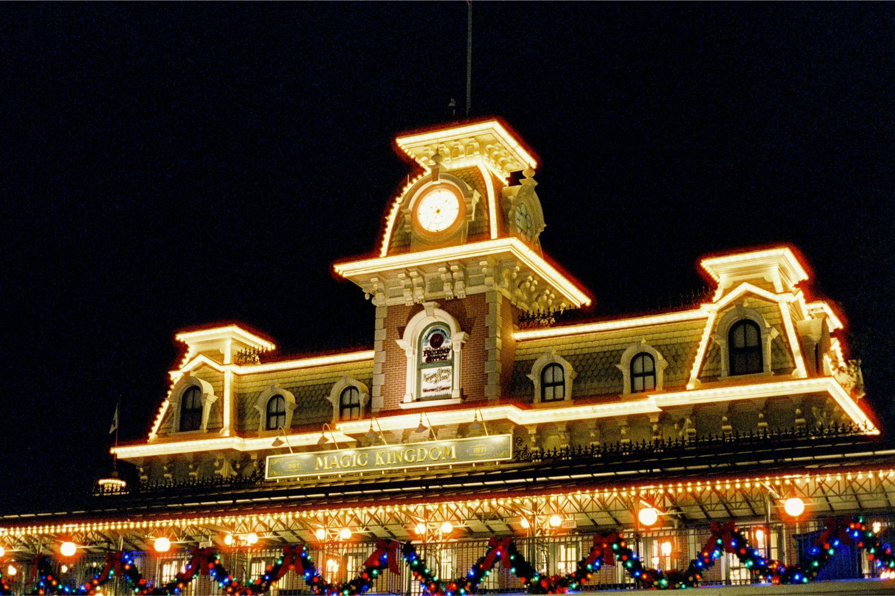 Magic Kingdom entrance station lit up with Christmas lights and holiday decorations at night.